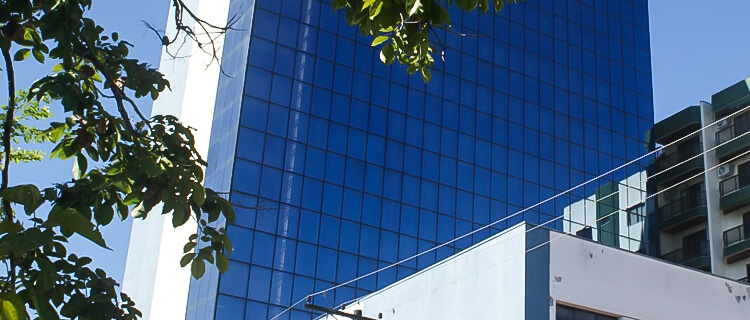 Prédio revestido de vidro azul. Ao lado esquerdo, há folhas verdes e ao lado direito, um prédio verde de menor altura.
